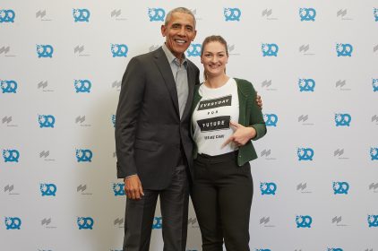 Digitalministerin Judith Gerlach, MdL, steht mit dem ehemaligen US-Präsidenten Barack Obama vor einer Pressewand, auf die blaue Brezen gedruckt sind. Sie trägt ein T-Shirt mit der Aufschrift: ‚Everyday for Future. Yes we can‘.