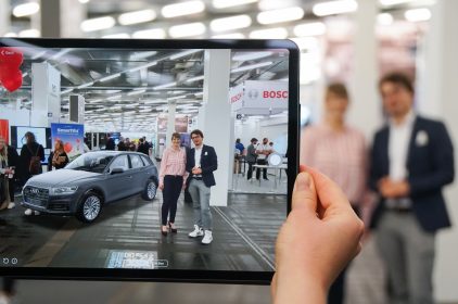 Das Bild zeigt ein Tablet, auf dessen Bildschirm Digitalministerin Judith Gerlach, MdL, und ein Besucher der AWE Europe zu sehen sind. Links auf dem Bidlschirm ist virtuell ein Auto in die Messehalle projiziert.