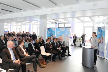 Digitalministerin Judith Gerlach, MdL, steht am Rednerpult bei dem Neujahrsempfang von M-net und haelt eine Rede. Links von ihr sind zahlreiche Gäste zu sehen.