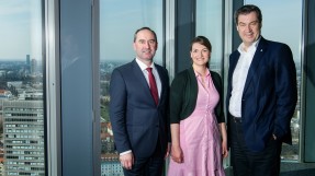 Digitalministerin Judith Gerlach, mit Ministerpräsident Dr. Markus Söder und Wirtschaftsminister Hubert Aiwanger stehen in einem Raum vor einer Fensterfront. Draußen ist die Stadt München zu erkennen.