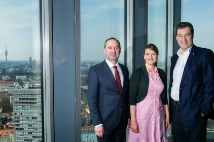 Digitalministerin Judith Gerlach, mit Ministerpräsident Dr. Markus Söder und Wirtschaftsminister Hubert Aiwanger stehen in einem Raum vor einer Fensterfront. Draußen ist die Stadt München zu erkennen.