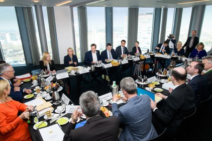 Die Mitglieder des Bayerischen Kabinetts sitzen an Konferenztischen in einem Raum eines Hochhauses. Durch die Fenster sind Häser zu sehen.