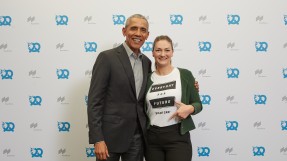 Digitalministerin Judith Gerlach, MdL, steht mit dem ehemaligen US-Präsidenten Barack Obama vor einer Pressewand, auf die blaue Brezen gedruckt sind. Sie trägt ein T-Shirt mit der Aufschrift: ‚Everyday for Future. Yes we can‘.