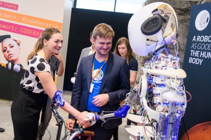 Digitalministerin Judith Gerlach, MdL, schüttelt dem humanoiden Roboter Roboy beim Bayerischen Digitalgipfel die Hand. Ein Mann steht daneben und lächelt. Foto: StMD / Jörg Koch