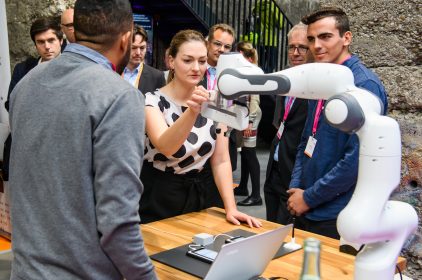 Digitalministerin Judith Gerlach, MdL, berührt einen Roboterarm. Teilnehmer des Digitalgipfels schauen ihr dabei zu. Foto: StMD / Jörg Koch