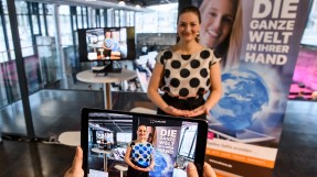 Staatsministerin Gerlach steht im Hintergrund. Im Vordergrund ist ein Livebild vom Tablet zu sehen, auf dem sie eine virtuelle Weltkugel in den Händen hält. Foto: StMD / Jörg Koch