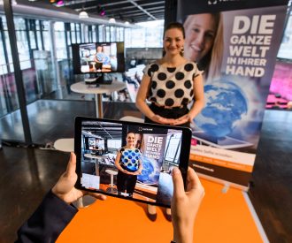 Staatsministerin Gerlach steht im Hintergrund. Im Vordergrund ist ein Livebild vom Tablet zu sehen, auf dem sie eine virtuelle Weltkugel in den Händen hält. Foto: StMD / Jörg Koch