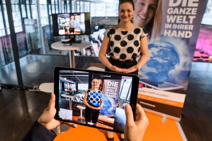 Staatsministerin Gerlach steht im Hintergrund. Im Vordergrund ist ein Livebild vom Tablet zu sehen, auf dem sie eine virtuelle Weltkugel in den Händen hält. Foto: StMD / Jörg Koch