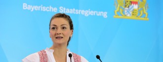 Digitalministerin Judith Gerlach steht im Dirndl hinter einem Rednerpult und spricht. Auf der blauen Wand hinter ihr steht ‚Bayerische Staatsregierung‘. Rechts daneben ist das große bayerische Staatswappen zu sehen.