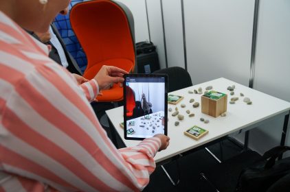 Digitalministerin Judith Gerlach, MdL, betrachtet durch ein Tablet Steine und Holzblöcke, die auf einem Tisch angeodnet sind. Im Tablet-Bildschirm ist eine Bekassine virtuell eingeblendet.