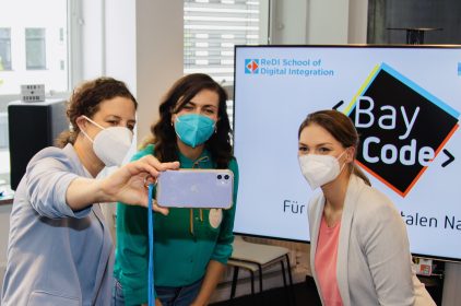 Birgit Köbl, Lena Rogl und Digitalministerin Gerlach stehen vor einem großen Bildschirm mit BayCode-Logo und machen ein Selfie