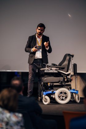 Ein Mann steht mit smarter Brille und Rollstuhl auf der Bühne.