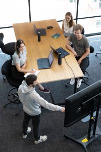 Eine Gruppe sitzt an einem Tisch und richtet ihre Aufmerksam auf den Referenten, der neben dem Großbildschirm steht.
