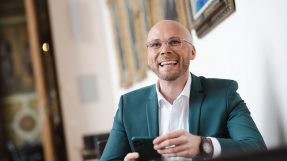 Staatsminister Dr. Fabian Mehring lacht in die Kamera und hält ein Smartphone in der Hand.