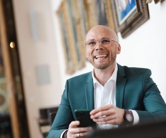Staatsminister Dr. Fabian Mehring lacht in die Kamera und hält ein Smartphone in der Hand.