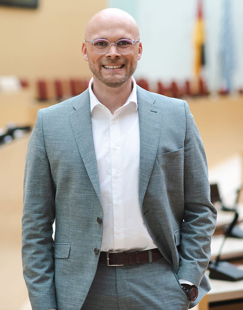 Staatsminister Dr. Fabian Mehring lächelt in die Kamera und steht im Bayerischen Landtag.
