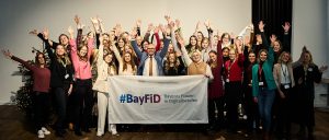 Gruppenbild von Teilnehmerinnen des BayFiD-Programms und Staatsminister Dr. Mehring. Die Peronen strecken die Hände in die Luft. In der ersten Reihe halten sie ein Banner mit der Aufschrift "BayFiD"