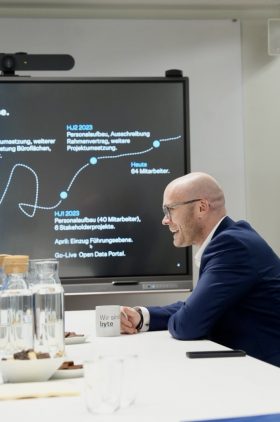 Digitalminister Dr. Fabian Mehring sitzt lachend an einem Tisch. Er hat eine Tasse mit der Aufschrift Wir sind BYTE in der Hand. Im Hintergrund ist ein Bildschirm mit einem Zeitstrahl zu sehen.