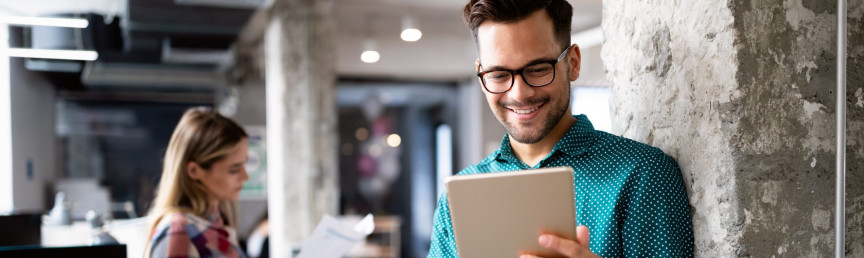 Mann mit Tablet in der Hand, im Hintergrund Frau mit Unterlagen in der Hand