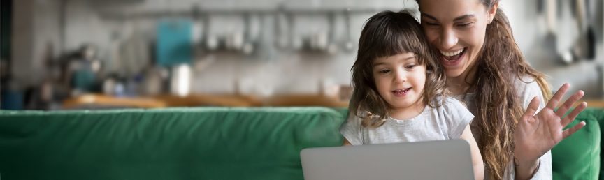 Eine Frau sitzt lachend mit einem Kind auf dem Schoß auf einem Sofa und winkt in die Kamera eines aufgeschlagenen Laptops.