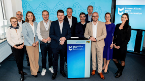 Gruppenfoto von elf Personen, die vor einem blauen Hintergrund mit dem Logo 'Bayern-Allianz gegen Desinformation' stehen. Die Gruppe besteht aus Männern und Frauen in formeller und halbformeller Kleidung, die sich um ein Rednerpult versammelt haben.