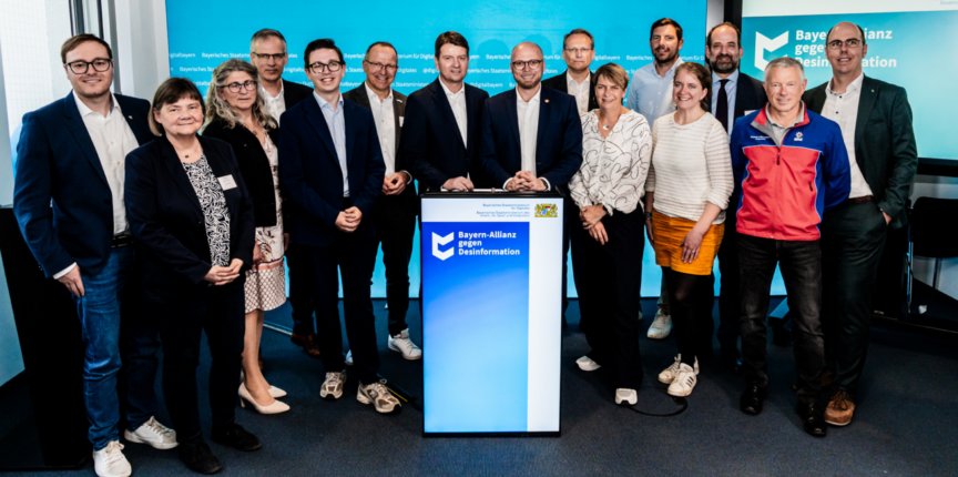 Eine Gruppe von Personen steht auf einer Bühne vor einem Podium mit der Aufschrift 'Bayern-Allianz gegen Desinformation'. Die Gruppe besteht aus Männern und Frauen, die sich um das Podium versammelt haben. Im Hintergrund ist ein Banner mit demselben Logo und Text zu sehen.
