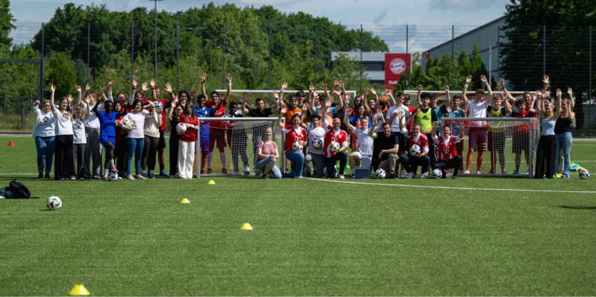 Eine Gruppe von Menschen steht auf einem Fußballfeld und posiert für ein Gruppenfoto. Viele von ihnen halten Fußballbälle und tragen verschiedene Sportkleidungen. Einige Personen knien oder hocken in der ersten Reihe, während andere stehen und ihre Hände heben. Im Hintergrund sind zwei kleine Fußballtore und ein Zaun zu sehen, hinter dem Bäume und ein Gebäude sichtbar sind.
