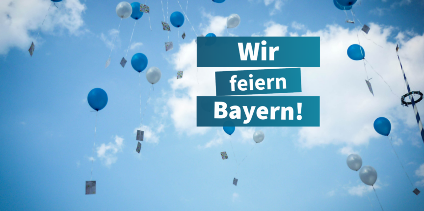 Ein klarer blauer Himmel mit vielen weißen und blauen Ballons, die in die Luft steigen. Im Vordergrund steht der Text 'Wir feiern Bayern!' in weißen Buchstaben auf grünem Hintergrund.