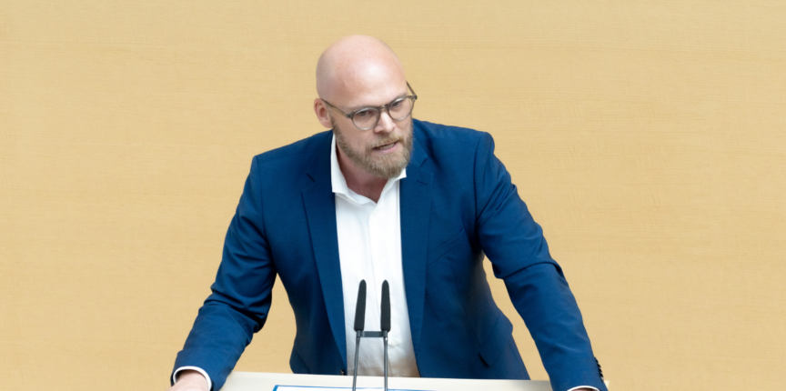 Ein Mann mit Brille und Bart steht an einem Pult mit Mikrofonen. Er trägt ein weißes Hemd und ein blaues Sakko. Der Hintergrund ist einfarbig beige.